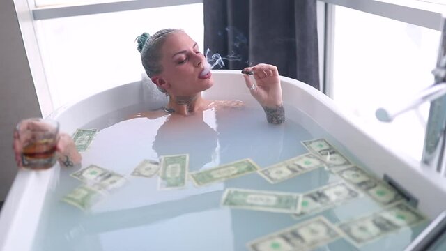 A young sexy tattooed girl enjoying a cigar and a drink while having a bath in a relaxed atmosphere in the bathroom with a bunch of money in the water. Relaxation, luxury, bathroom, home