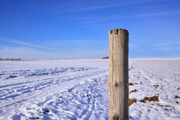 A weathered fence post stands in a wide winter landscape in Bavaria, against snow and a blue sky