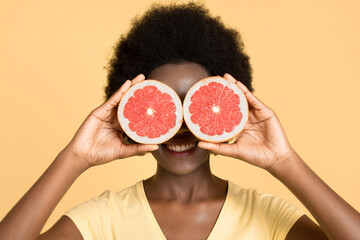 Close up horizontal shot of playful african american woman smiling while posing with two grapefruit slices covering her eyes, isolated over yellow wall background