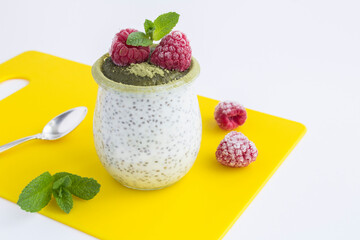 Chia pudding with raspberries and matcha tea in the glass jar on the yellow cutting board on the white background