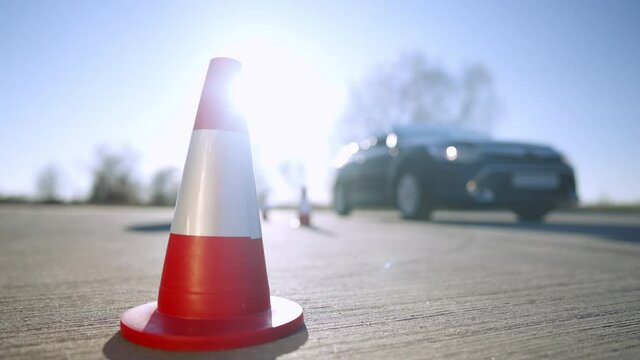 Close-up of road cone outdoors in sunlight with blurred driving instructor walking to car at background. Professional man on autumn or spring day. Driving school training.