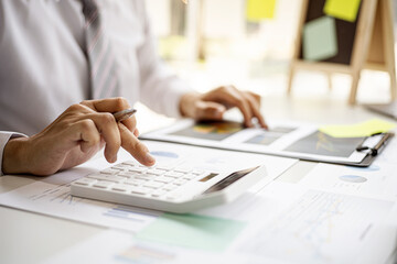 A business man is using a calculator to calculate company financial figures from earnings papers, a businessman sitting in his office where the company financial chart is placed. Financial concept.