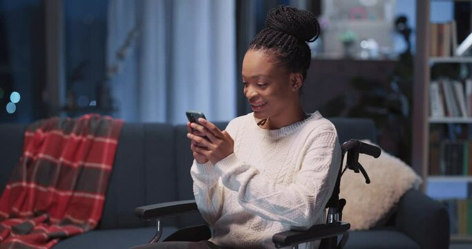 Positive afro-american casual girl using mobile phone receiving text messages rejoicing. Black disabled young woman in wheelchair.