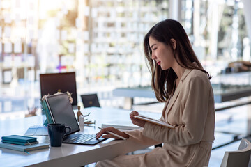 Young Asian woman working on a tablet and taking notes at a modern office.