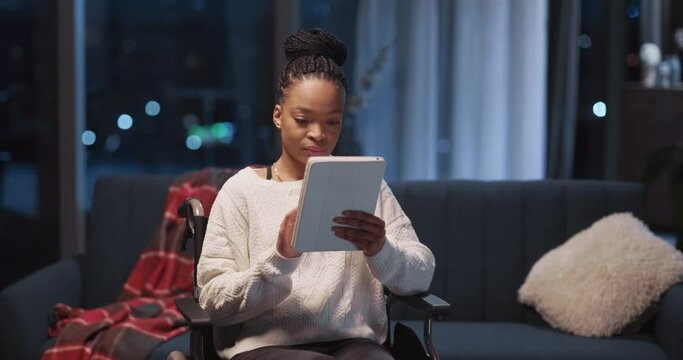 Beautiful cheerful african young woman using tablet computer apps playing mobile game sitting in wheelchair inside home apartment.