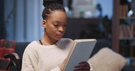 Young pretty african serious woman sitting in wheelchair browsing internet news on mobile tablet computer digital device at home. - Powered by Adobe