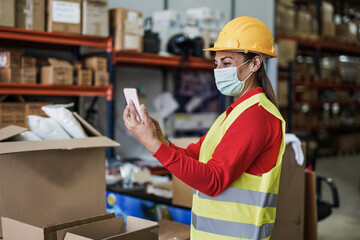 Mature latin woman with smartphone working inside warehouse while wearing safety face mask for coronavirus outbreak