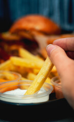 Young girl hand holding a dipped French fries into white sauce with onion rings and chicken stripes in the back