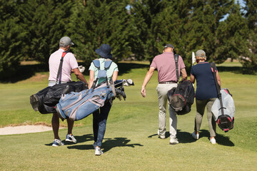 Four caucasian senior men and women wearing face masks walking across golf course holding golf bags