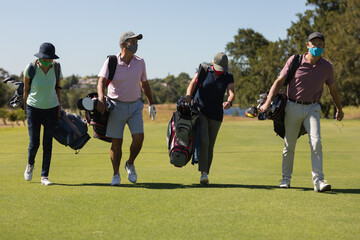 Four caucasian senior men and women wearing face masks walking across golf course holding golf bags
