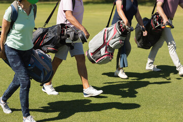 Four caucasian senior men and women wearing face masks walking across golf course holding golf bags