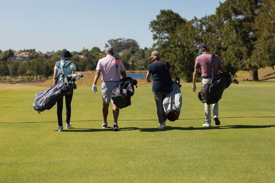 Four caucasian senior men and women wearing face masks walking across golf course holding golf bags - Powered by Adobe