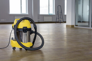 Yellow vacuum cleaner on the wooden floor against the background of an empty office building after renovation.  The concept of cleaning premises after renovation. Copy space.