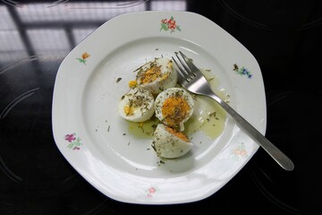 Hard-boiled chicken eggs with oregano, olive oil, black pepper and a fork on a white ceramic plate on a kitchen hob. Top view, selective focus