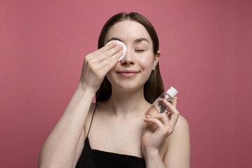 Smiling young woman with closed eyes removing makeup with cotton pad, holding bottle of micellar water on pink background. Girl cleaning face. Treatment and cosmetology. Beauty routine, skincare