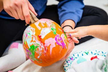 An unrecognizable child finger painting a pumpkin with colorful paints while an adult holds it.