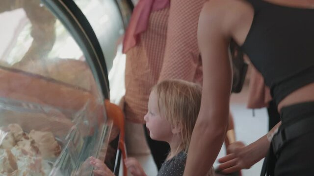 Young Caucasian woman wearing casual clothes standing at showcase in cafe, holding her toddler daughter who is choosing ice-cream