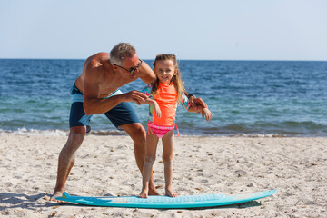 Fototapeta premium papà con occhiali da sole in costume da bagno insegna le basi del surf alla figlia in una spiaggia al mare..