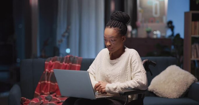 Young cheerful pretty african woman in wheelchair calling best friend on laptop webcam video conference communicating online inside apartment.