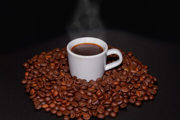 White coffee mug with a coffee drink inside and surrounded by roasted coffee beans of the Robusta variety.