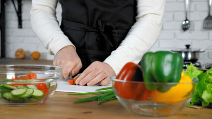 Professional chef cook man in black apron standing near table sliced fresh cherry tomato on cutting board at home kitchen. Middle shot male hands food vegetables ingredients for salad dish