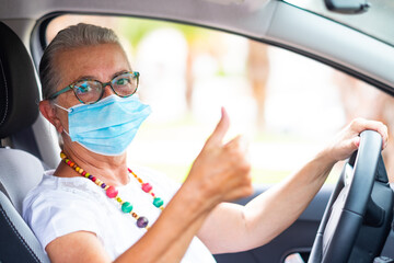 Coronavirus. Senior woman driving her car wearing surgical mask, looking at camera with thumb up. Focus on the face.