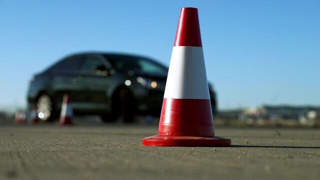 Close-up of red and white road cone with black car passing barriers. Unknown learner studying driving outdoors on sunny spring or auutmn day. Transportation and education concept.