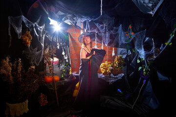 Blonde witch in red dress and black hat in Halloween decoration indoors.