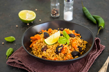Bulgur pilaf with carrots and black beans in a black plate on a dark concrete background. Vegetarian recipes.