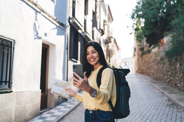 Joyful female tourist enjoying solo vacations using location map during travel sightseeing in touristic village, happy hipster girl connecting to roaming internet for making online video vlog