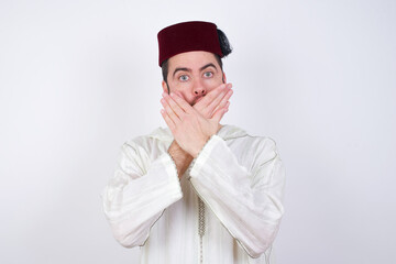 young handsome Caucasian man wearing Arab djellaba and Fez hat over white wall shocked covering mouth with hands for mistake. Secret concept.