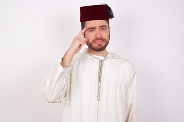 young handsome Caucasian man wearing Arab djellaba and Fez hat over white wall tries to memorize something, keeps fore finger on temple, reminds information for exam,