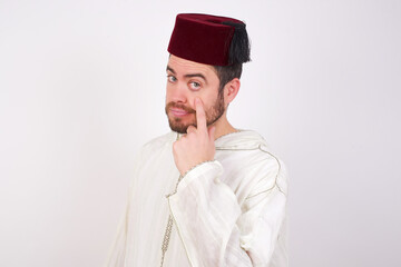 young handsome Caucasian man wearing Arab djellaba and Fez hat over white wall Pointing to the eye watching you gesture, suspicious expression.