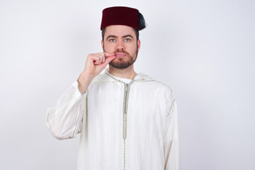 young handsome Caucasian man wearing Arab djellaba and Fez hat over white wall mouth and lips shut as zip with fingers. Secret and silent, taboo talking.
