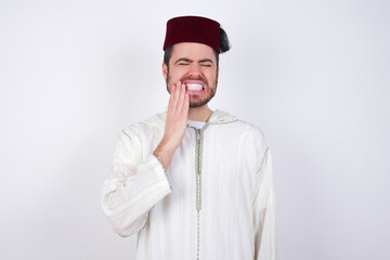 young handsome Caucasian man wearing Arab djellaba and Fez hat over white wall touching mouth with hand with painful expression because of toothache or dental illness on teeth.