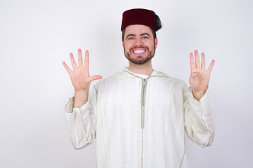 young handsome Caucasian man wearing Arab djellaba and Fez hat over white wall showing and pointing up with fingers number nine while smiling confident and happy.