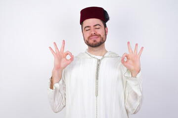 young handsome Caucasian man wearing Arab djellaba and Fez hat over white wall relax and smiling with eyes closed doing meditation gesture with fingers. Yoga concept.
