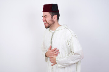 young handsome Caucasian man wearing Arab djellaba and Fez hat over white wall with hand on stomach because nausea, painful disease feeling unwell. Ache concept.