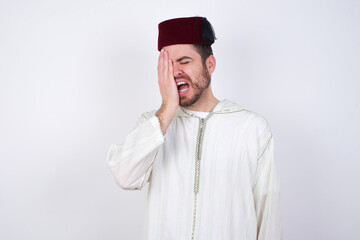 young handsome Caucasian man wearing Arab djellaba and Fez hat over white wall Yawning tired covering half face, eye and mouth with hand. Face hurts in pain.