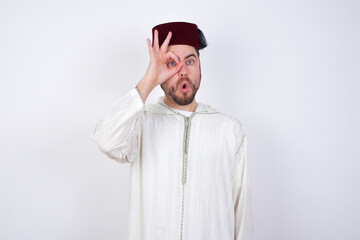 young handsome Caucasian man wearing Arab djellaba and Fez hat over white wall doing ok gesture shocked with surprised face, eye looking through fingers. Unbelieving expression.