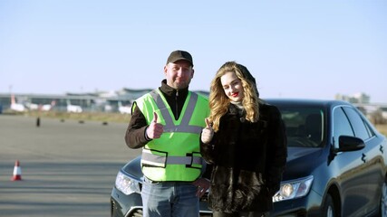 Positive male driving instructor and female student showing thumbs up looking at camera smiling. Portrait of happy young beautiful Caucasian woman passing test for driving license outdoors.
