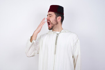 young handsome Caucasian man wearing Arab djellaba and Fez hat over white wall being tired and yawning after spending all day at work.