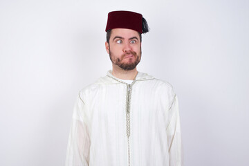young handsome Caucasian man wearing Arab djellaba and Fez hat over white wall making grimace and crazy face, screaming out of control, funny lunatic expressing freedom and wild.