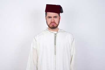 Displeased upset young handsome Caucasian man wearing Arab djellaba and Fez hat frowns face as going to cry, being discontent and unhappy as can't achieve goals,  Disappointed model has troubles