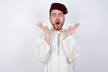 Surprised terrified young handsome Caucasian man wearing Arab djellaba and Fez hat over white wall...