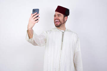 young handsome Caucasian man wearing Arab djellaba and Fez hat over white wall smiling and taking a...