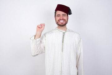 young handsome Caucasian man wearing Arab djellaba and Fez hat over white wall pointing up with fingers number ten in Chinese sign language Shi