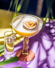 yellow tropical cocktail with passion fruit and shot on a background of tropical plant leaves and sun shades. Color background