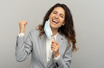 Studio portrait of happy business woman taking off mask, raising fist, grey background. Female removing medical mask from face and looking at camera. Coronavirus, Covid-19 ended. Pandemic is over.
