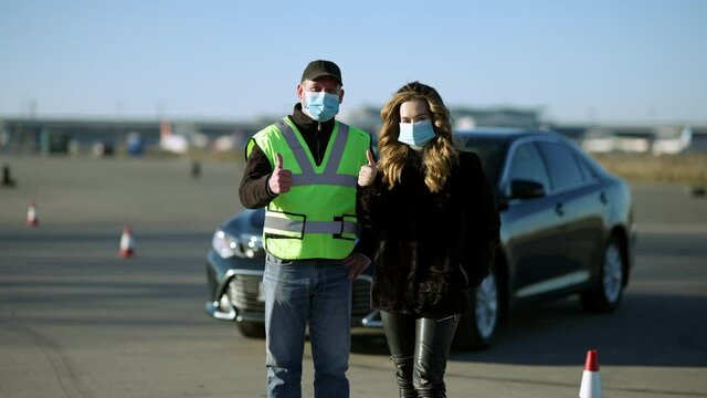 Confident Caucasian woman and man in Covid face masks showing thumbs up with car at background. Portrait of driving instructor and student posing on coronavirus pandemic outdoors.
